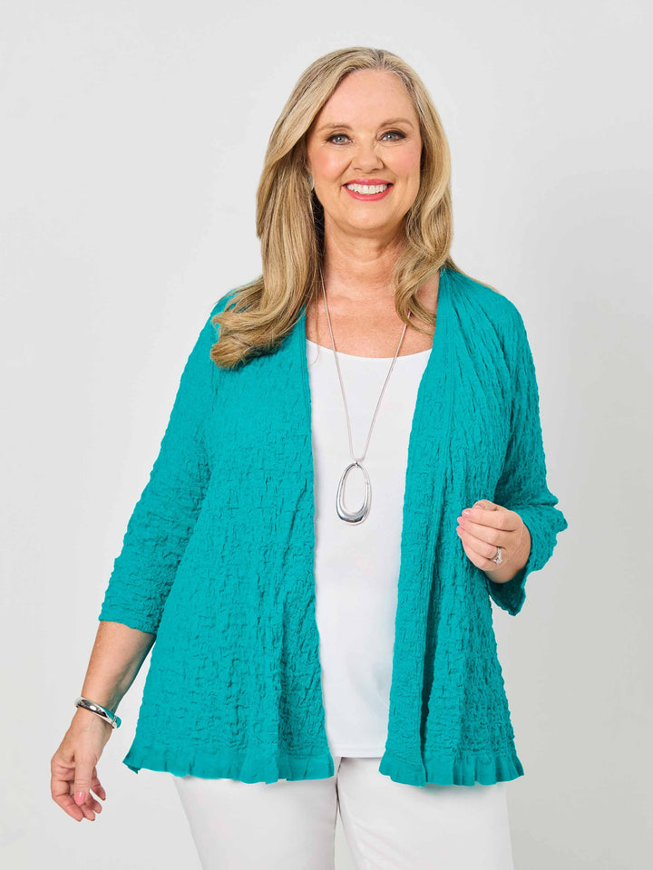 A woman wearing a textured teal open-front cardigan over a white top and white pants