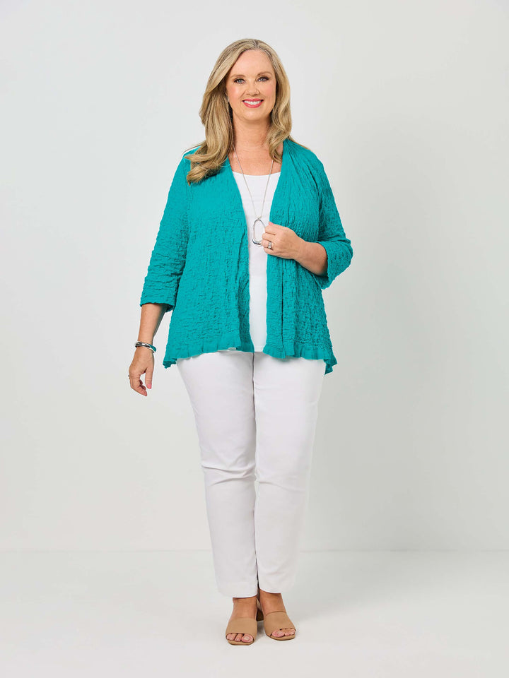 A woman wearing a textured teal open-front cardigan over a white top and white pants