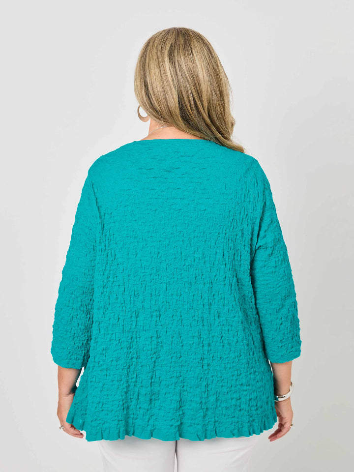 A woman wearing a textured teal open-front cardigan over a white top and white pants