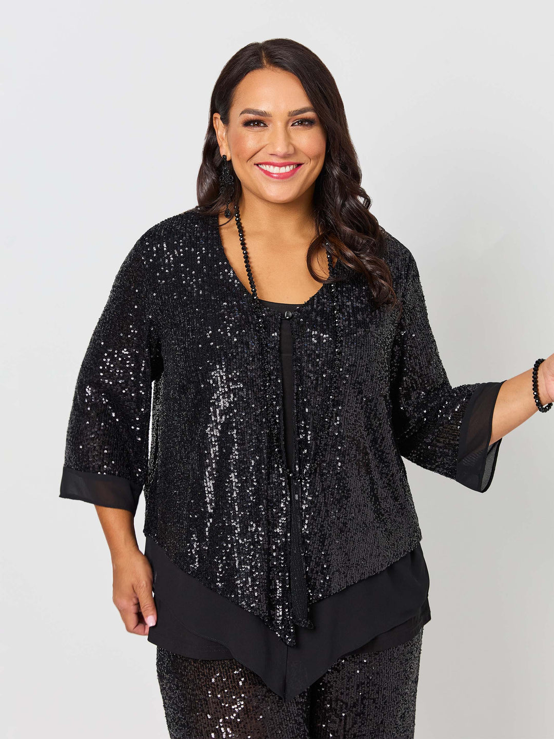 A woman wearing a black sequined three-quarter sleeve evening jacket with layered ruffle hem, paired with matching sequined pants