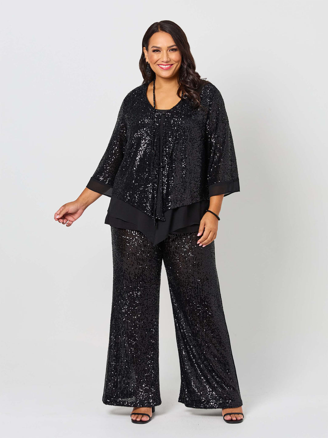 A woman wearing a black sequined three-quarter sleeve evening jacket with layered ruffle hem, paired with matching sequined pants
