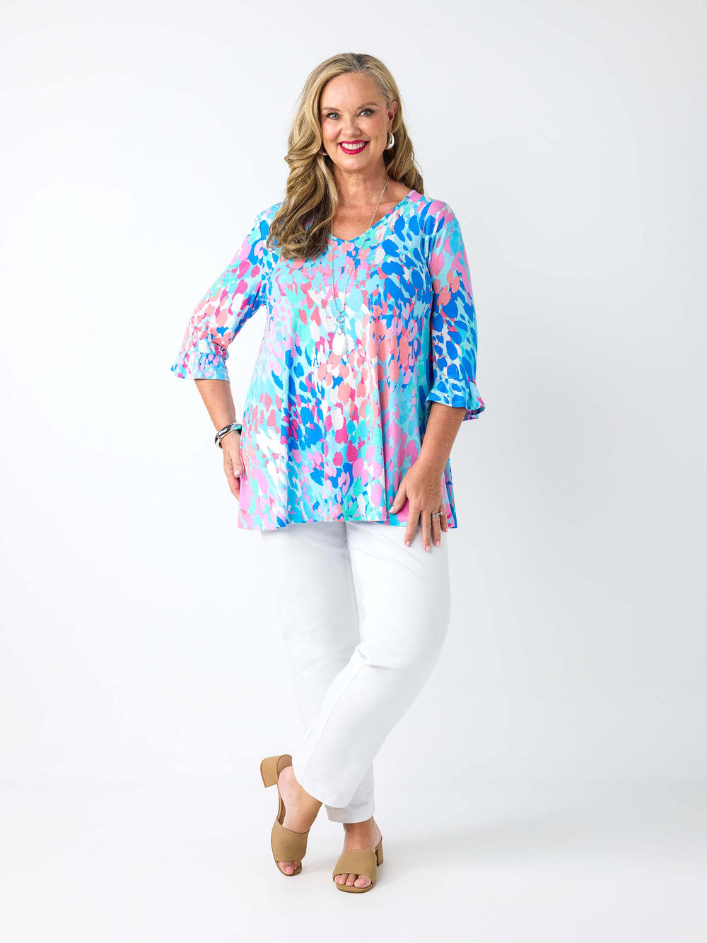 A woman wearing a v-neck top with 3 quarter frill sleeves in aqua blue and pink print, paired with white pant