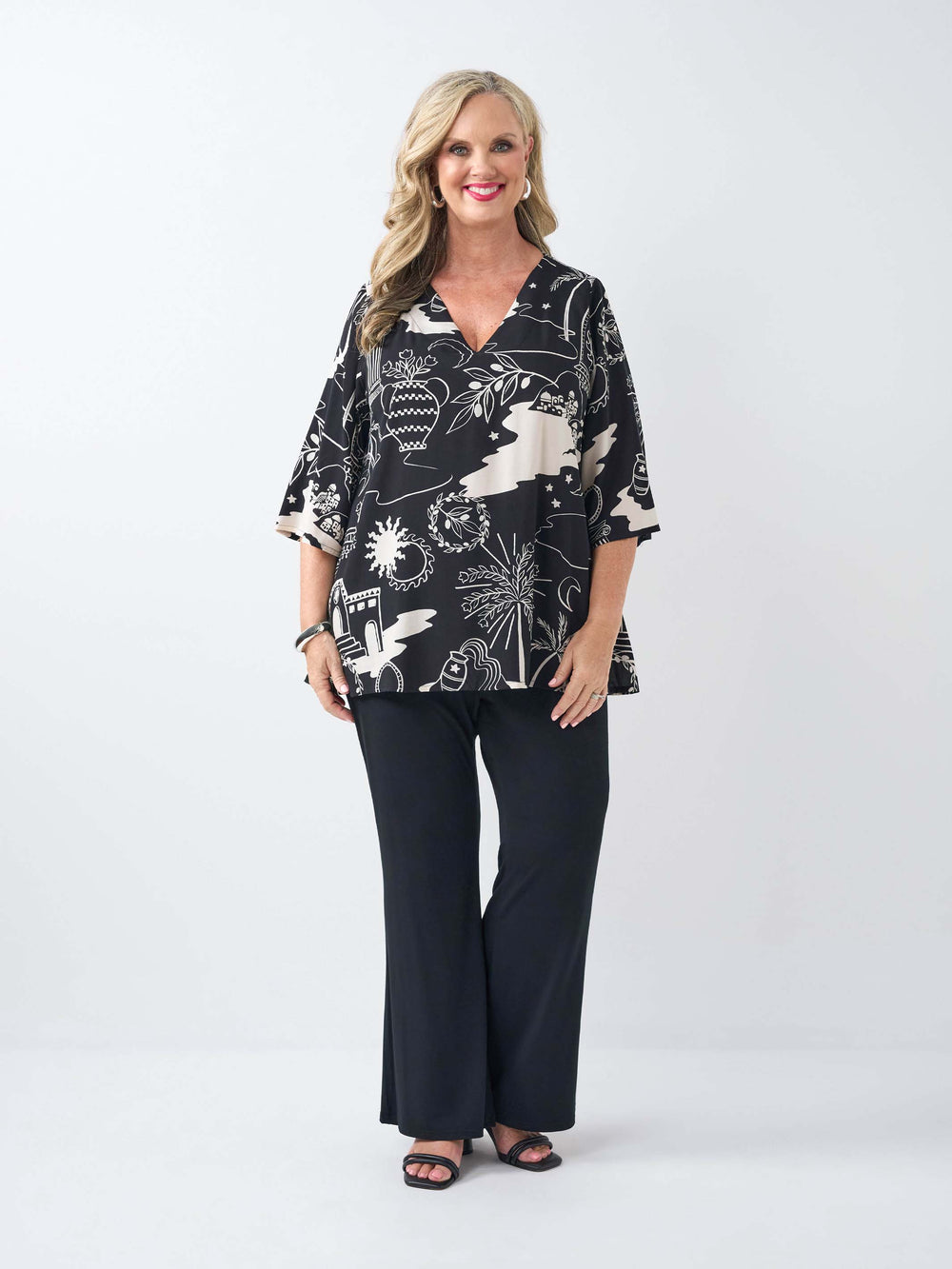 A woman wearing a black and cream abstract roman styled patterned v-neck top with three-quarter sleeves and black pants.