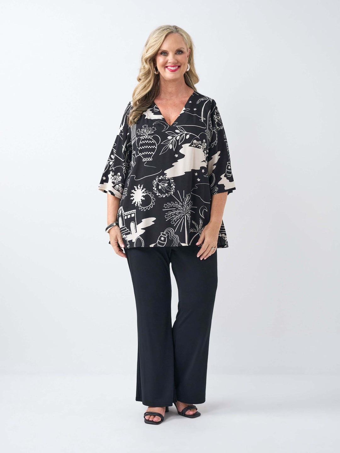 A woman wearing a black and cream abstract roman styled patterned v-neck top with three-quarter sleeves and black pants.