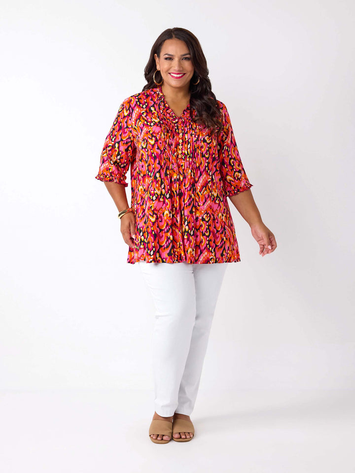 A woman wearing a vibrant, multicolour abstract print top with three-quarter ruffled sleeves and a smocked neckline, paired with white pants