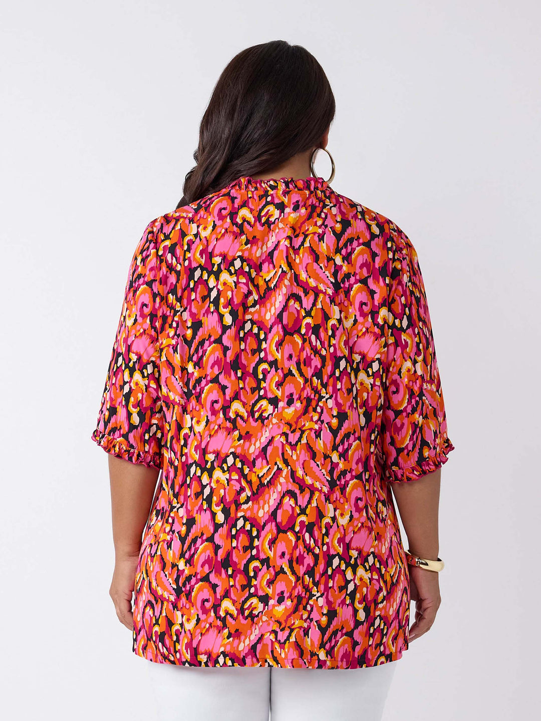 A woman wearing a vibrant, multicolour abstract print top with three-quarter ruffled sleeves and a smocked neckline, paired with white pants
