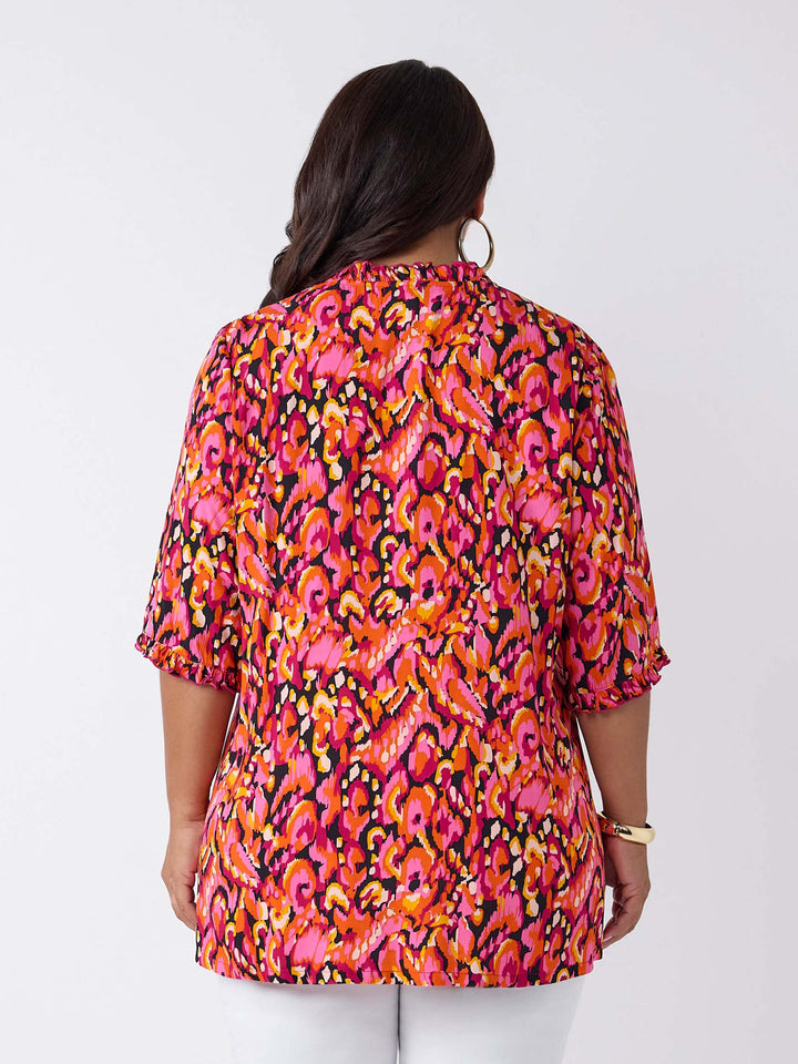 A woman wearing a vibrant, multicolour abstract print top with three-quarter ruffled sleeves and a smocked neckline, paired with white pants