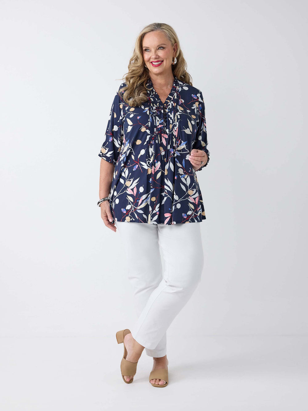 A woman wearing a navy blue leaf print v-neck top with white, pink, and beige botanical motifs, paired with white pants. The top has pintuck detail at the centre front and ruffle trim around the neckline and sleeves.