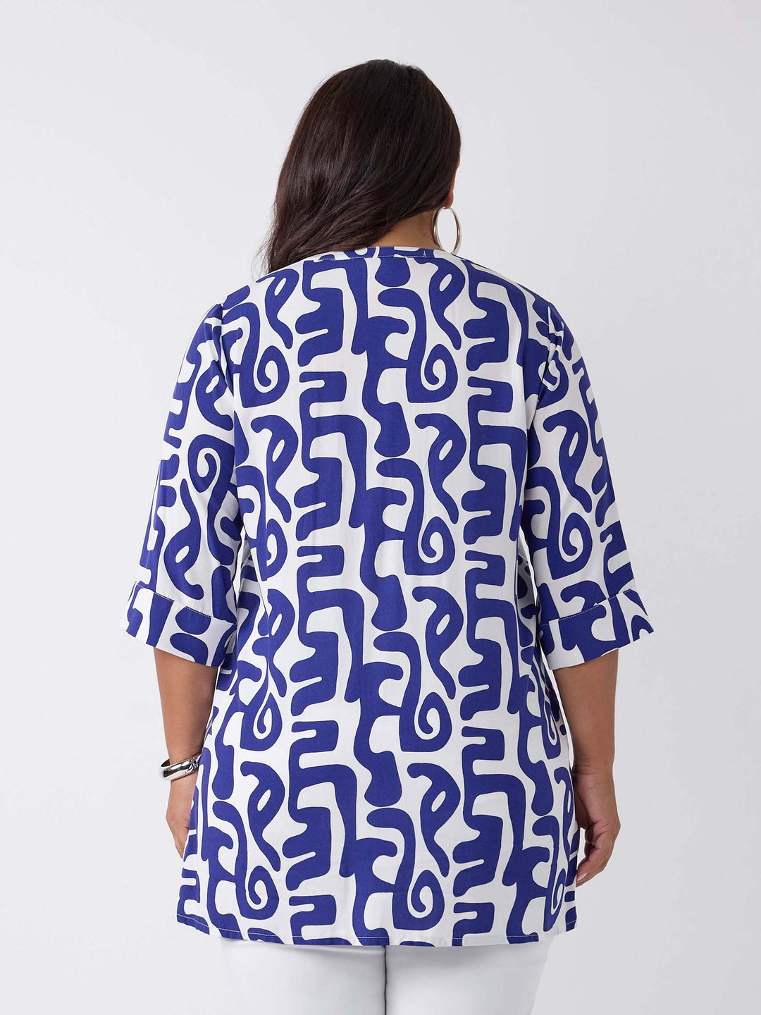 A woman wearing a blue and white abstract patterned v-neck blouse with three-quarter sleeves, paired with white pants
