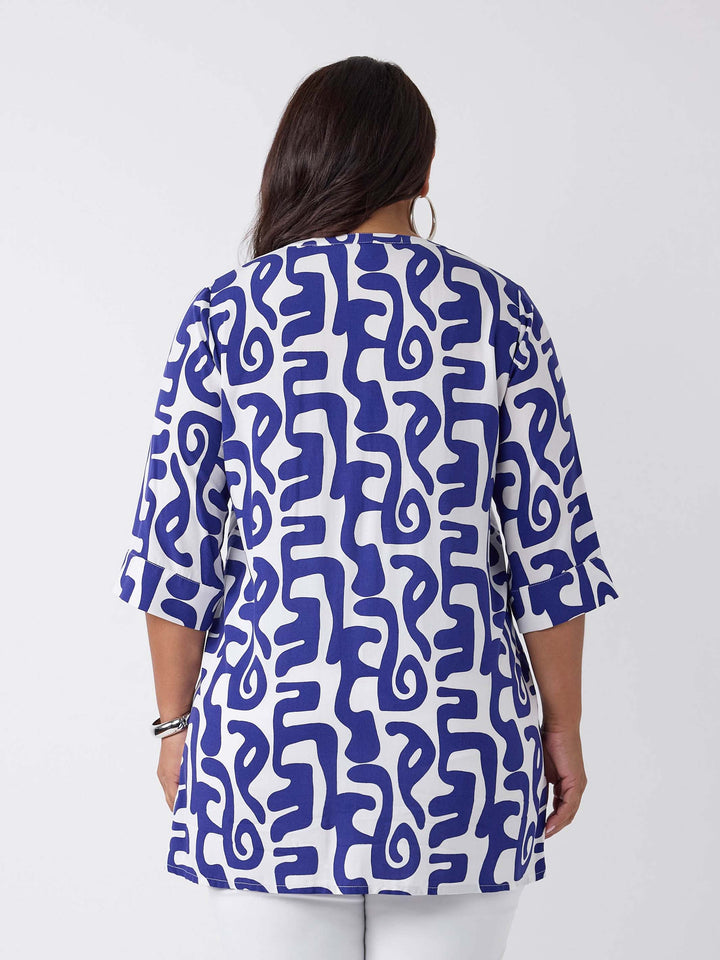 A woman wearing a blue and white abstract patterned v-neck blouse with three-quarter sleeves, paired with white pants