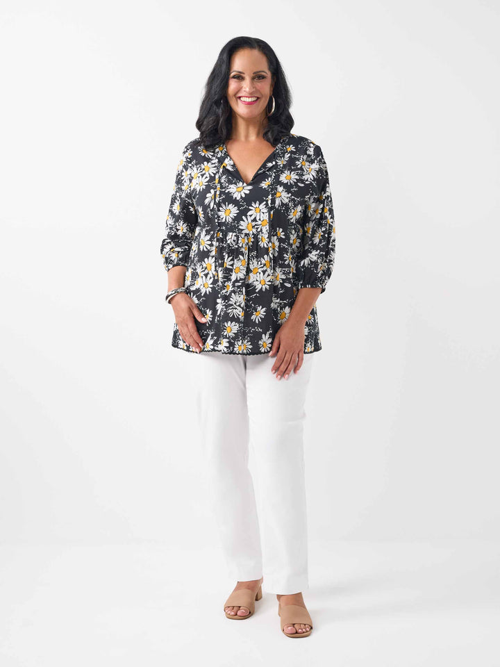 A woman wearing a black long-sleeve blouse with a white and yellow daisy floral print, paired with white pants