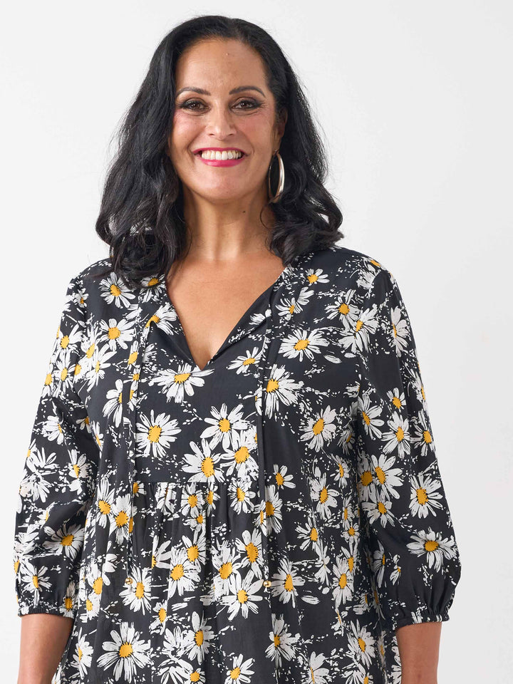 A detail view of a woman wearing a black long-sleeve blouse with a white and yellow daisy floral print, paired with white pants