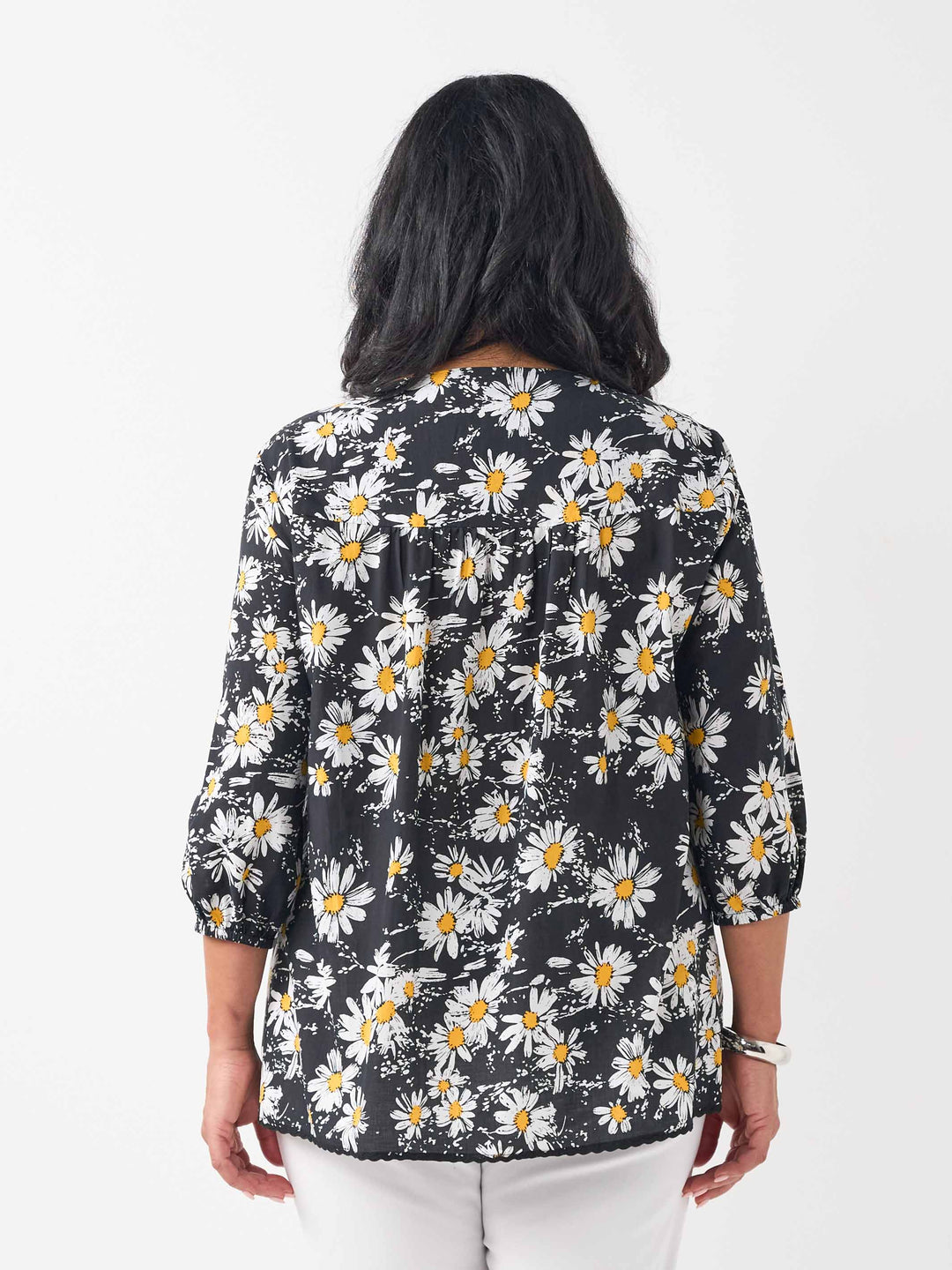 A  back view of a woman wearing a black long-sleeve blouse with a white and yellow daisy floral print, paired with white pants