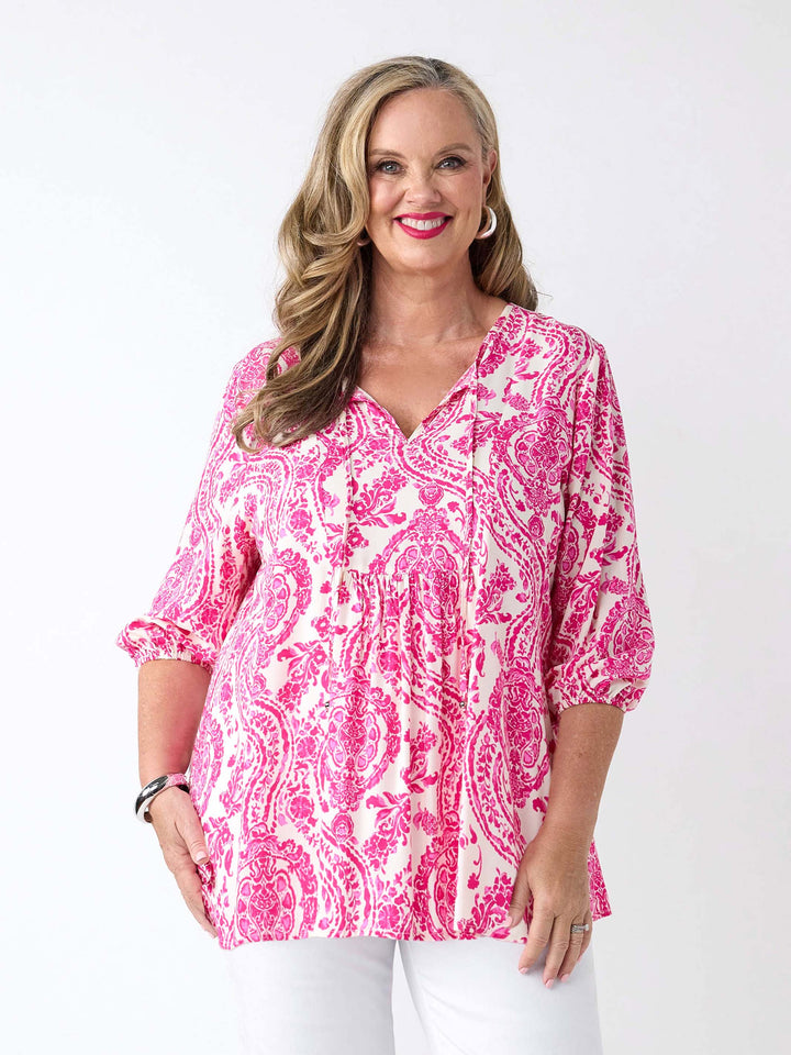 A woman wearing a pink and white paisley patterned blouse with v-neck tie front and three-quarter sleeves, paired with white pants
