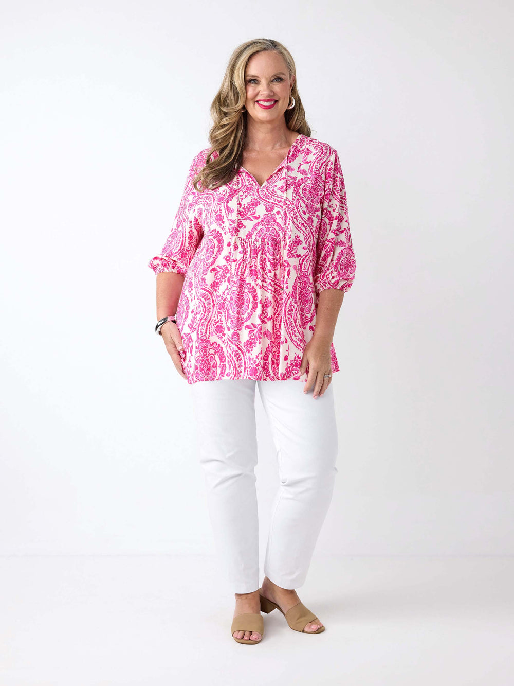 A woman wearing a pink and white paisley patterned blouse with v-neck tie front and three-quarter sleeves, paired with white pants