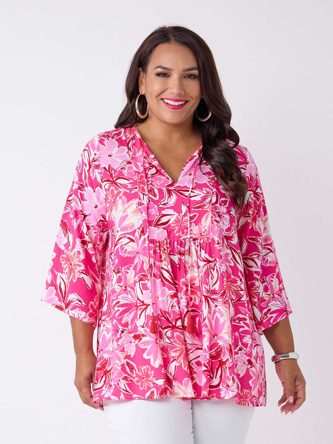 A woman wearing a v-neck top with tie neck detail in pretty bright pink floral print, paired with white pant