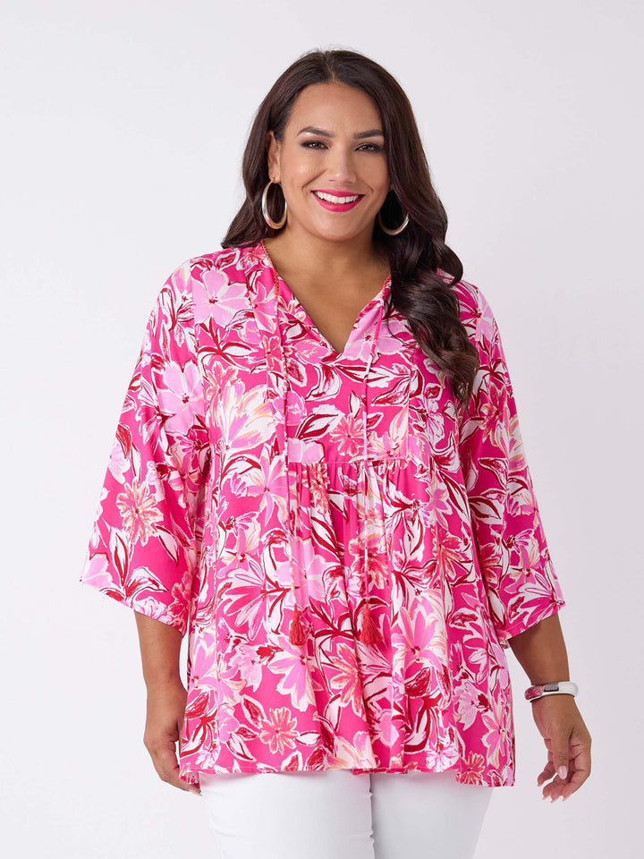 A woman wearing a v-neck top with tie neck detail in pretty bright pink floral print, paired with white pant