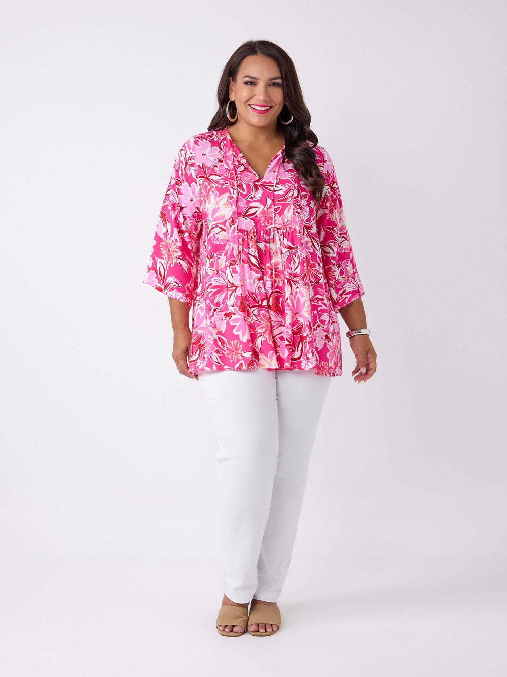 A woman wearing a v-neck top with tie neck detail in pretty bright pink floral print, paired with white pant