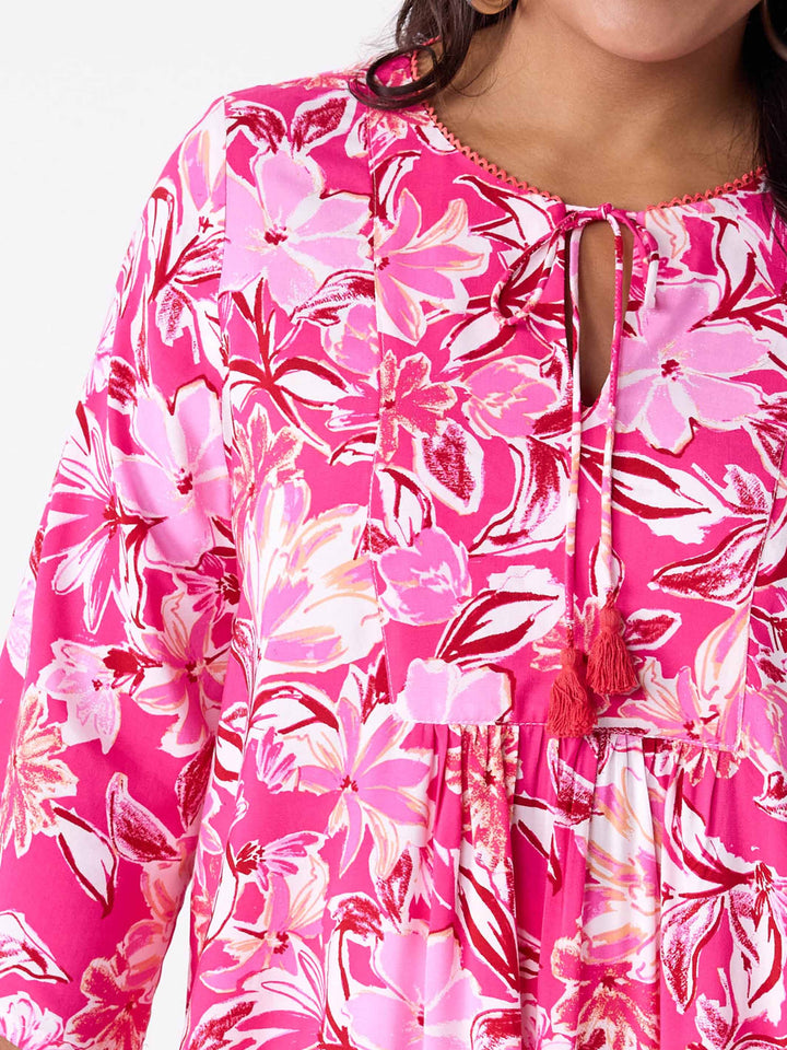 A woman wearing a v-neck top with tie neck detail in pretty bright pink floral print, paired with white pant