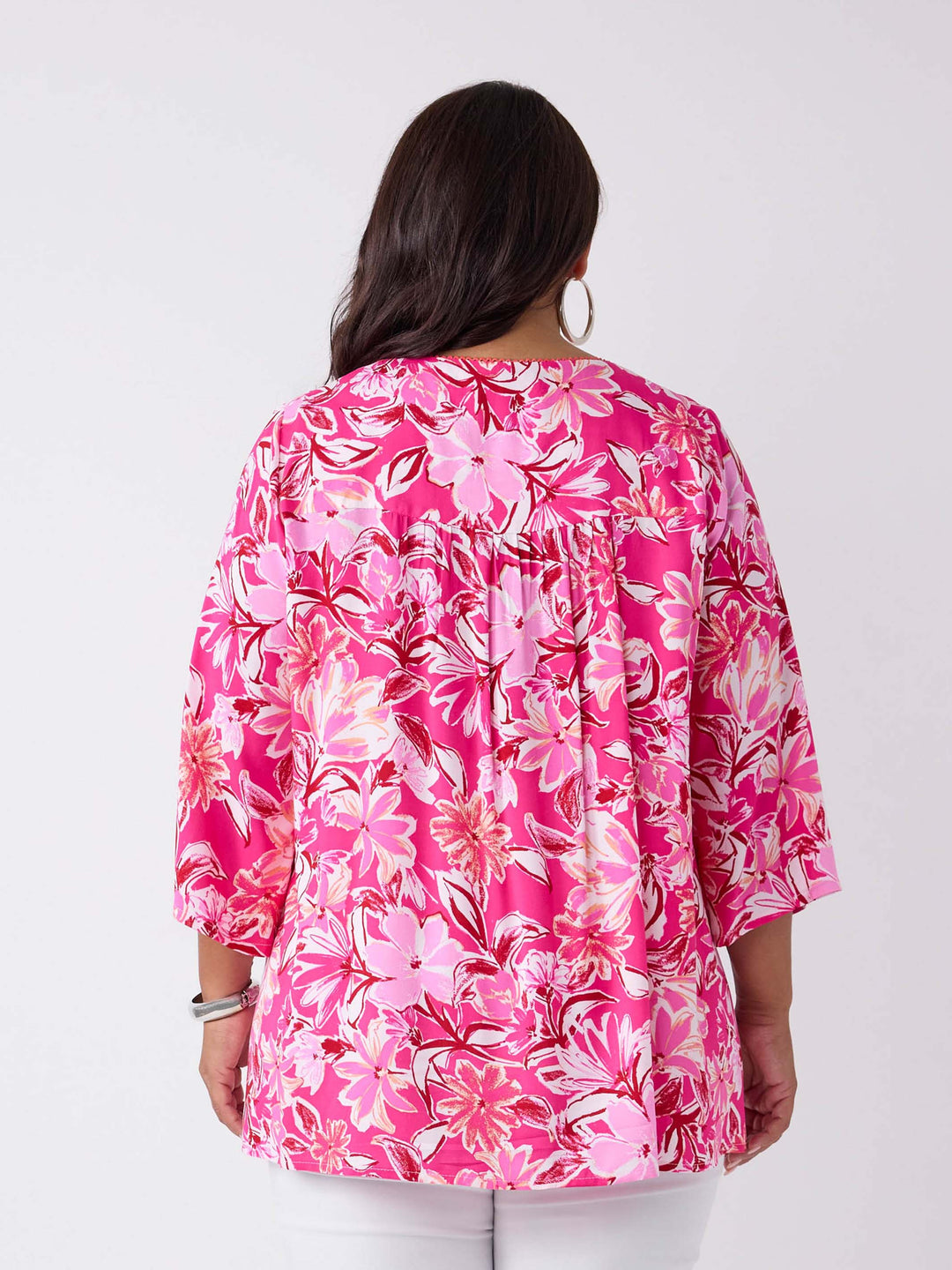 A woman viewed from the back wearing a v-neck top with tie neck detail in pretty bright pink floral print, paired with white pant