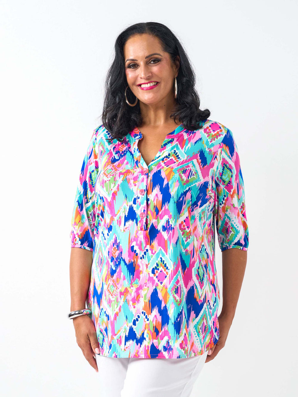 A woman wearing a vibrant, multicoloured short-sleeve button-up top with a colourful ikat-style geometric pattern in pink, blue, teal, and yellow, paired with white pants