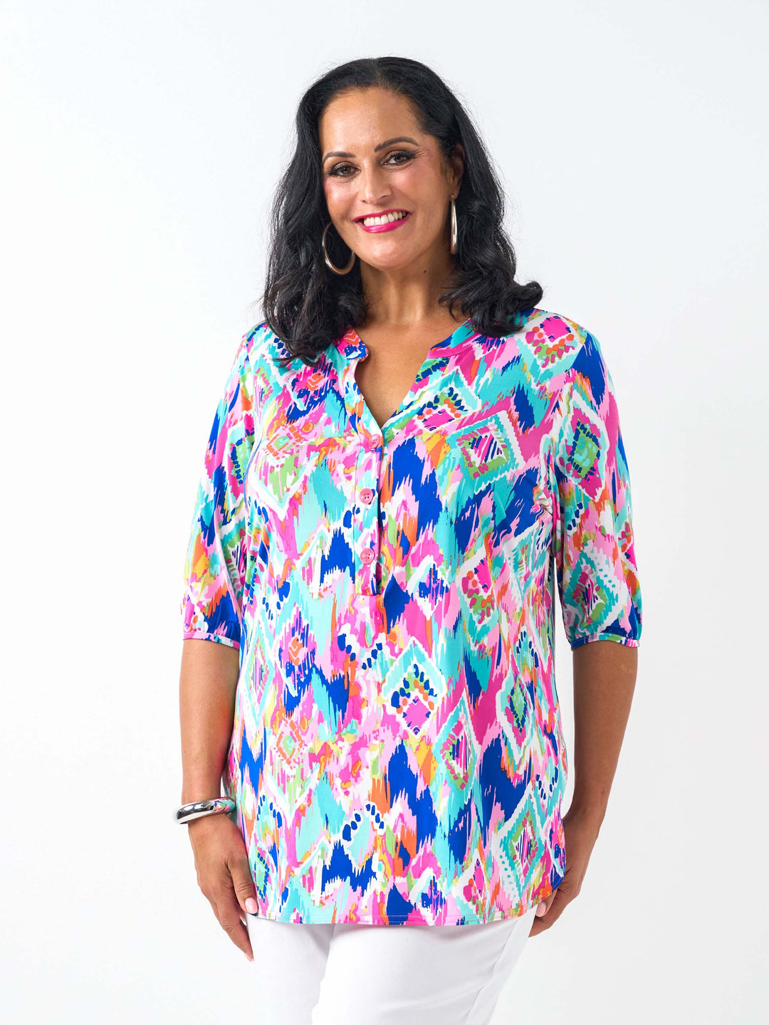A woman wearing a vibrant, multicoloured short-sleeve button-up top with a colourful ikat-style geometric pattern in pink, blue, teal, and yellow, paired with white pants