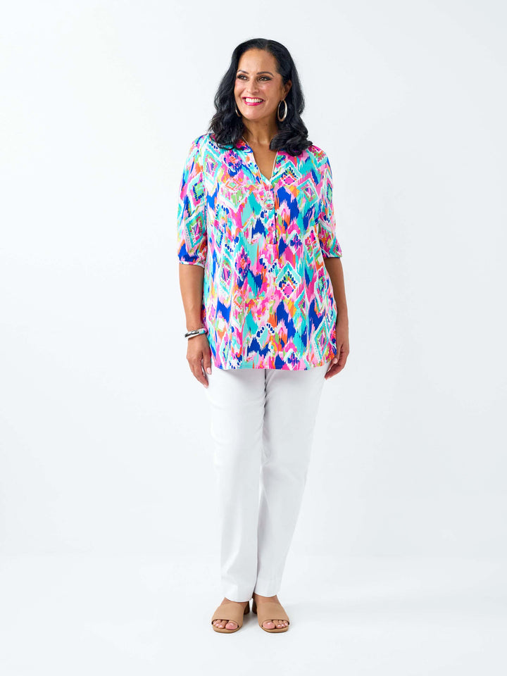 A woman wearing a short-sleeve button-up shirt with a vibrant multicolour geometric pattern, paired with white pants