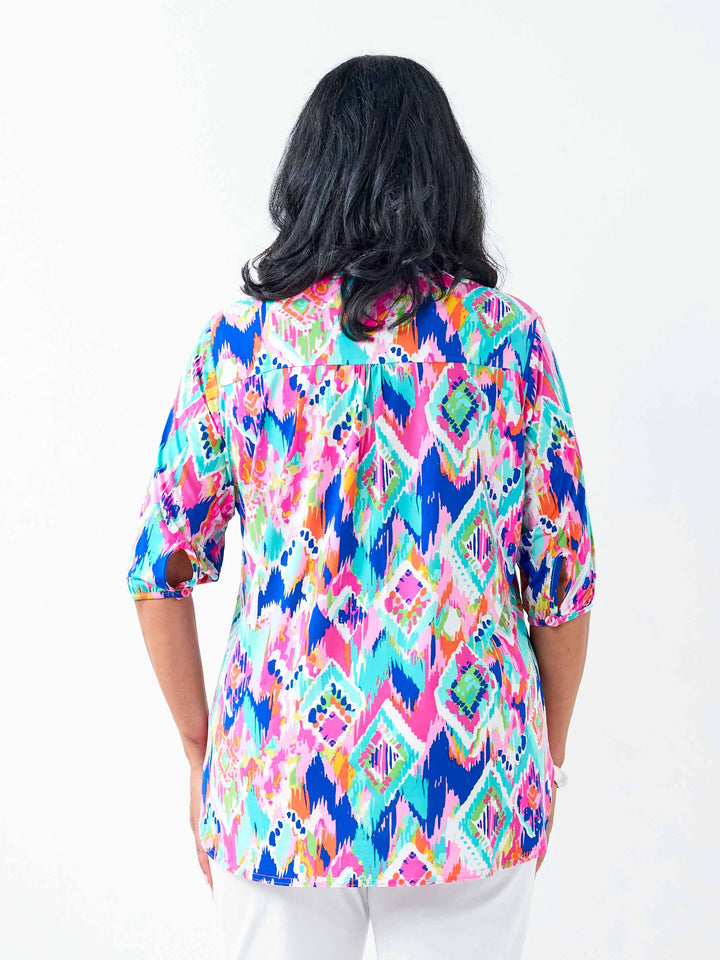 A woman viewed from behind wearing a vibrant, multicoloured geometric patterned button-up shirt with white pants