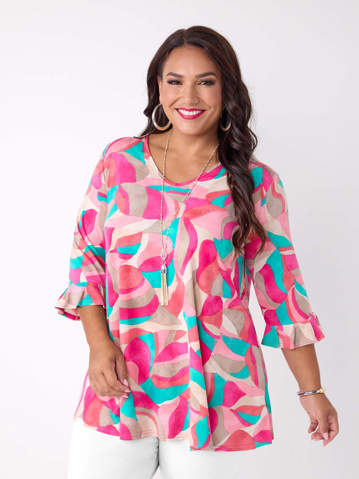 A woman wearing a pink, teal, and beige abstract patterned top top with three-quarter ruffled sleeves, paired with white pants