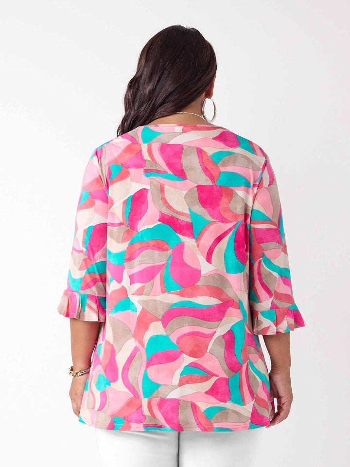 A woman viewed from the back wearing a pink, teal, and beige abstract patterned top top with three-quarter ruffled sleeves, paired with white pants