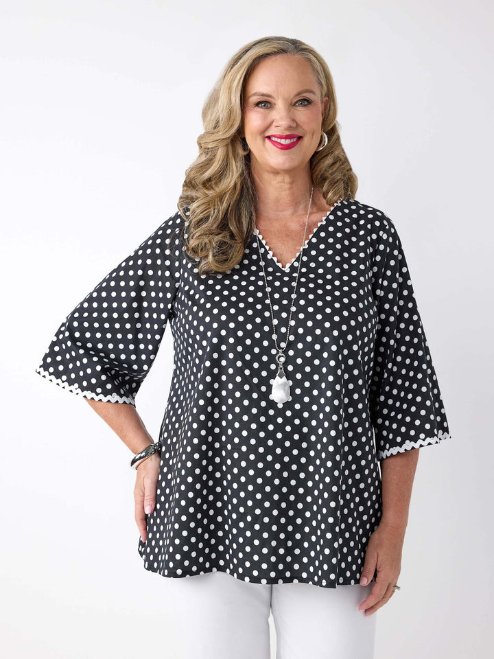 A woman wearing a black and white polka dot v-neck top with ric-rac lace detail around the neckline and sleeves 