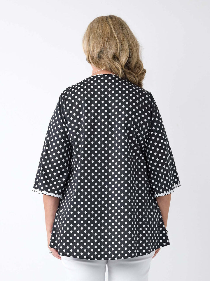 A woman viewed from the back wearing a black and white polka dot v-neck top with ric-rac lace detail around the neckline and sleeves 
