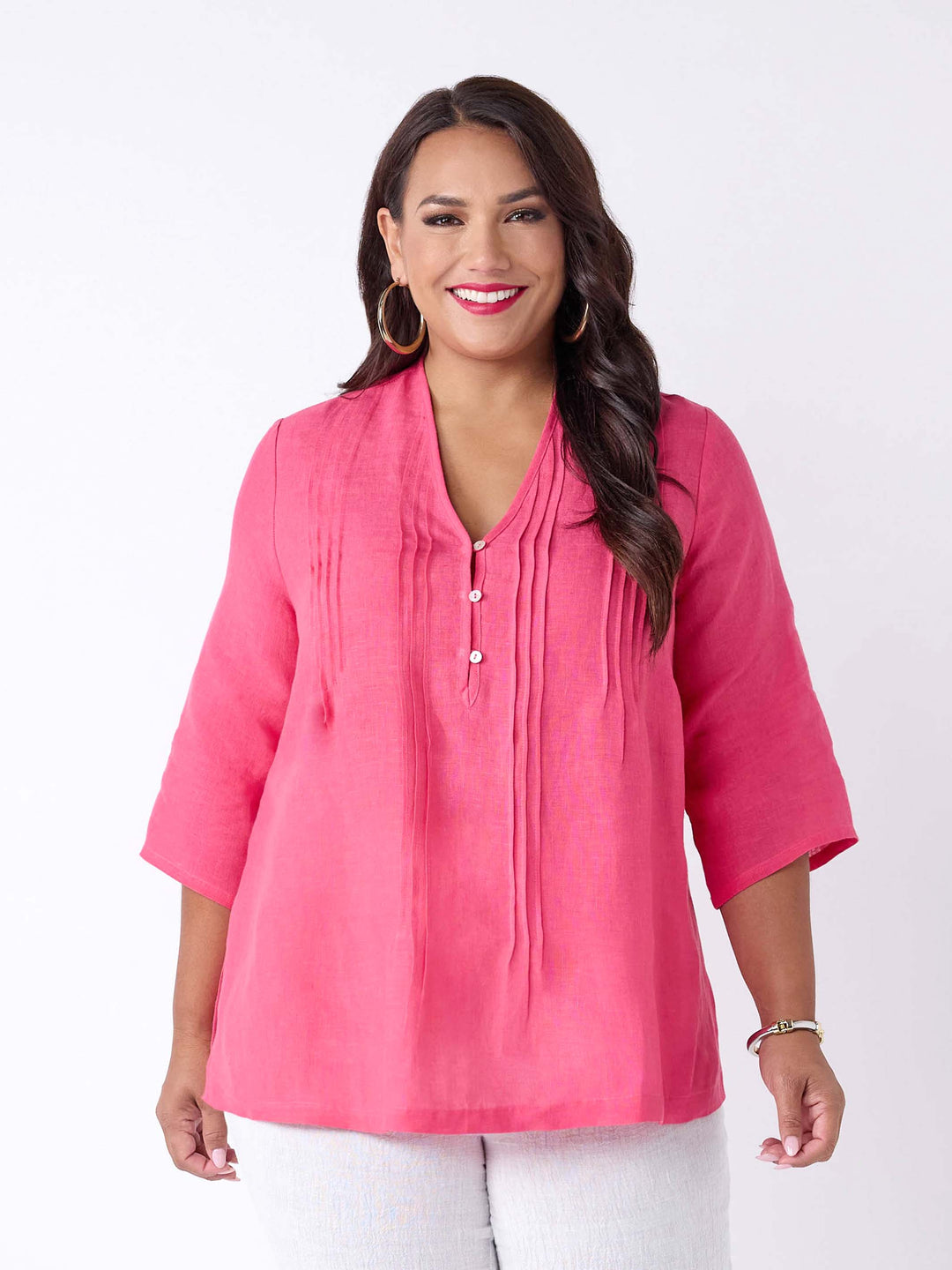 A woman wearing a bright pink linen three-quarter sleeve blouse with a pleated front and button placket, paired with white pants