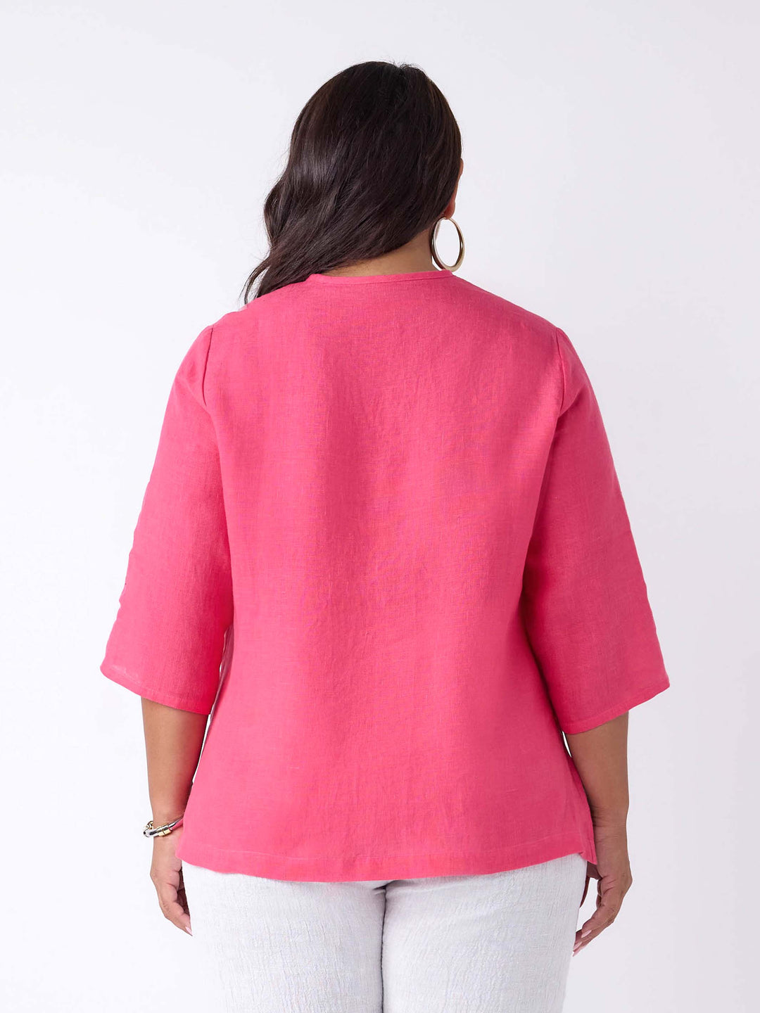 A woman wearing a bright pink linen three-quarter sleeve blouse with a pleated front and button placket, paired with white pants