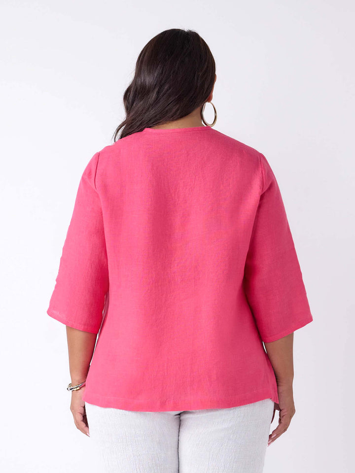 A woman wearing a bright pink linen three-quarter sleeve blouse with a pleated front and button placket, paired with white pants