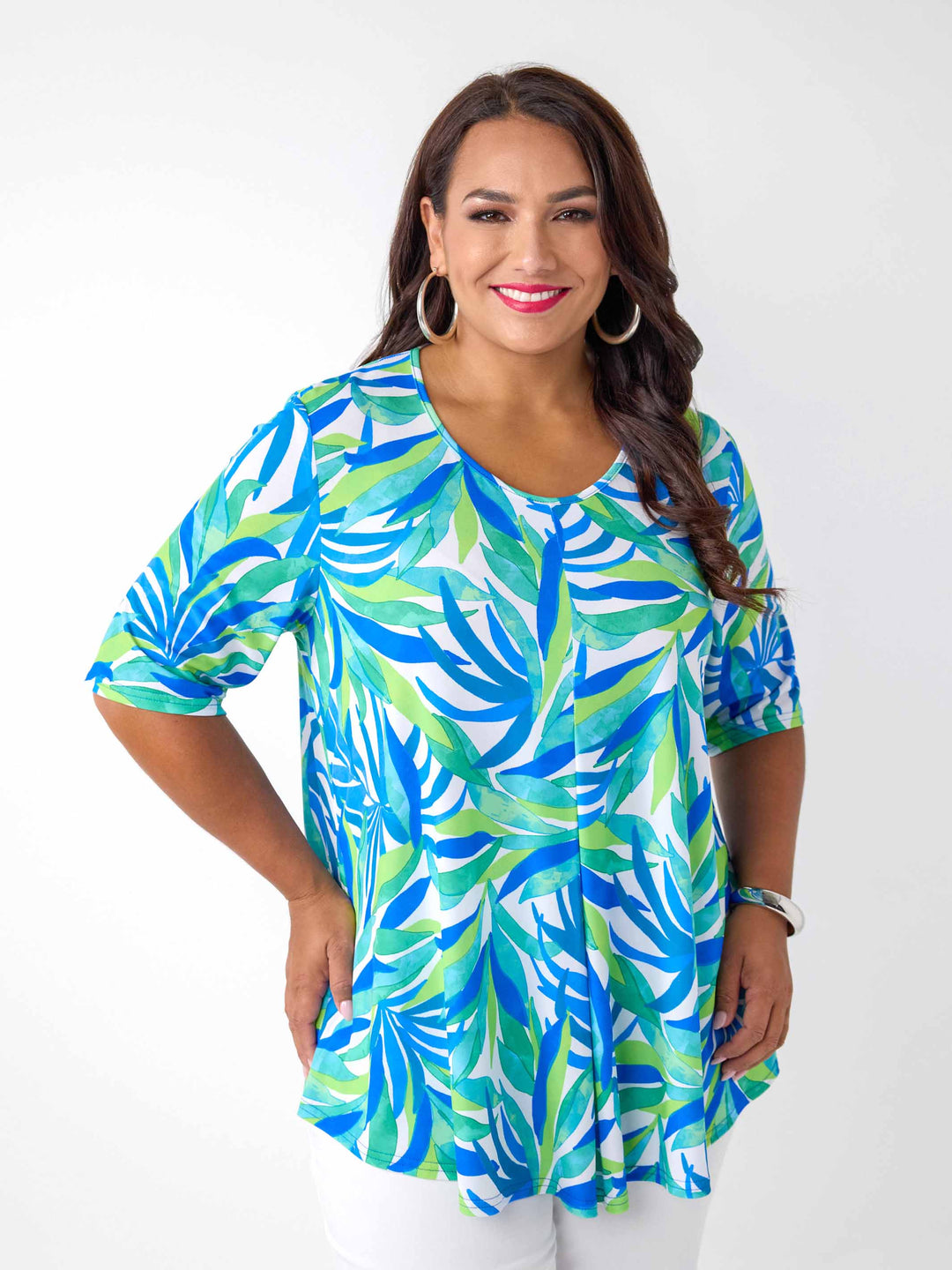 A woman wearing a short-sleeve v-neck top with a vibrant blue, green, and white tropical leaf pattern, paired with white pants