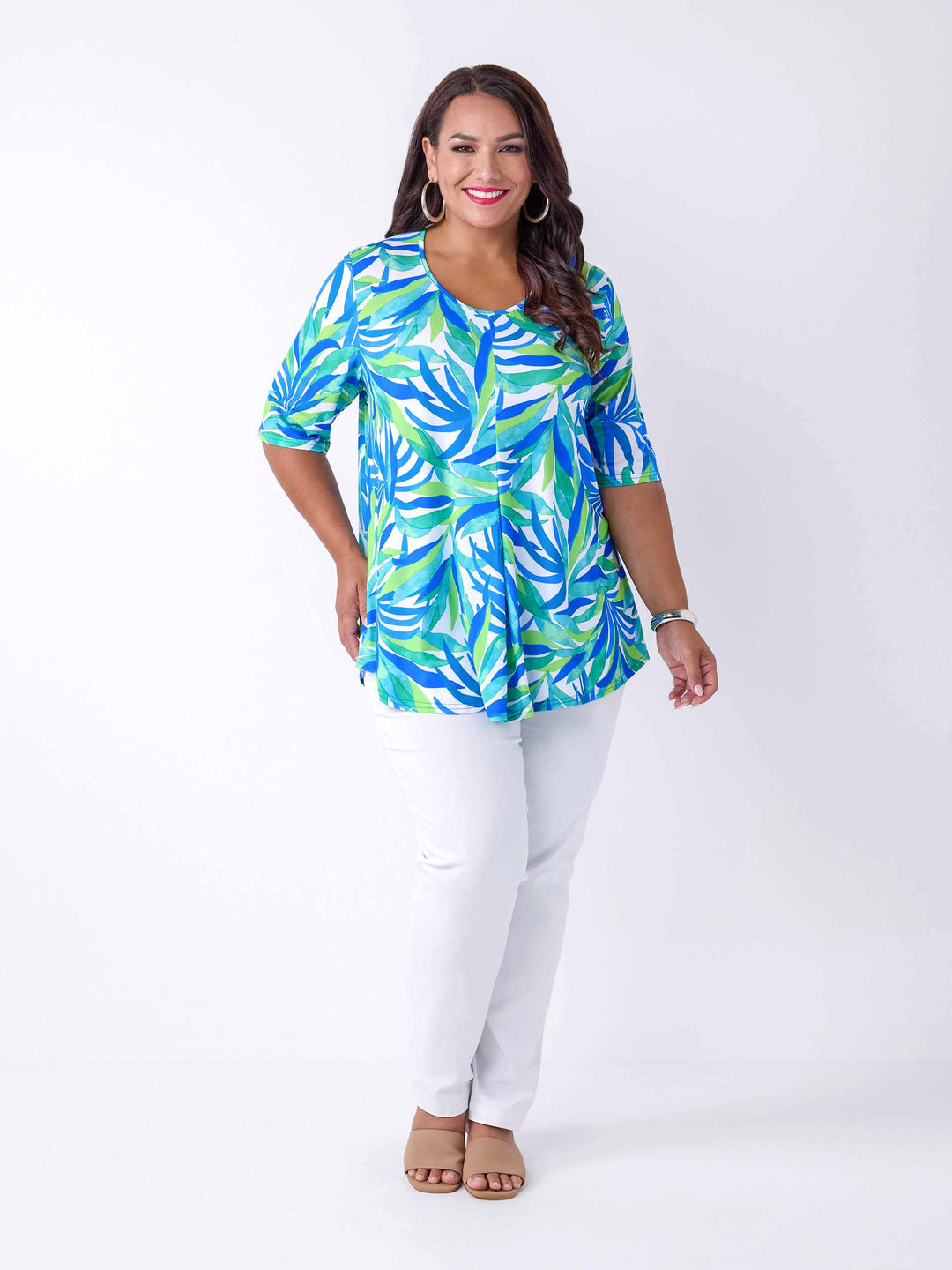 A woman wearing a short-sleeve v-neck top with a vibrant blue, green, and white tropical leaf pattern, paired with white pants