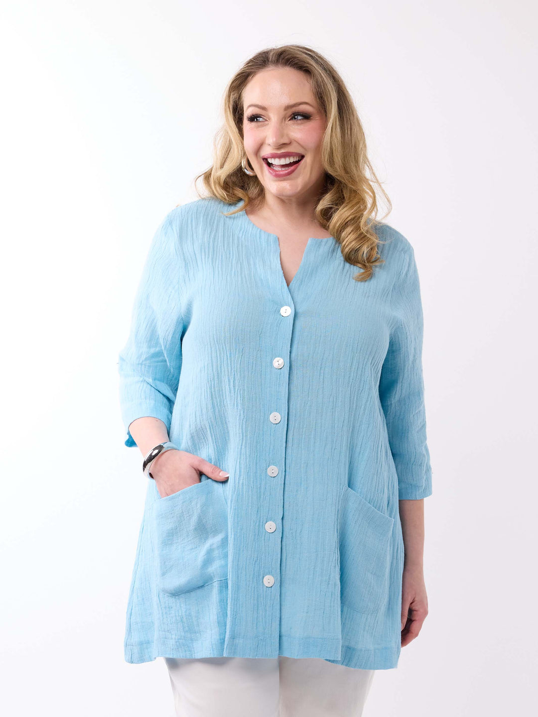 A woman wearing a light aqua blue crinkle-textured button-up top with white buttons and side pockets, paired with white pants