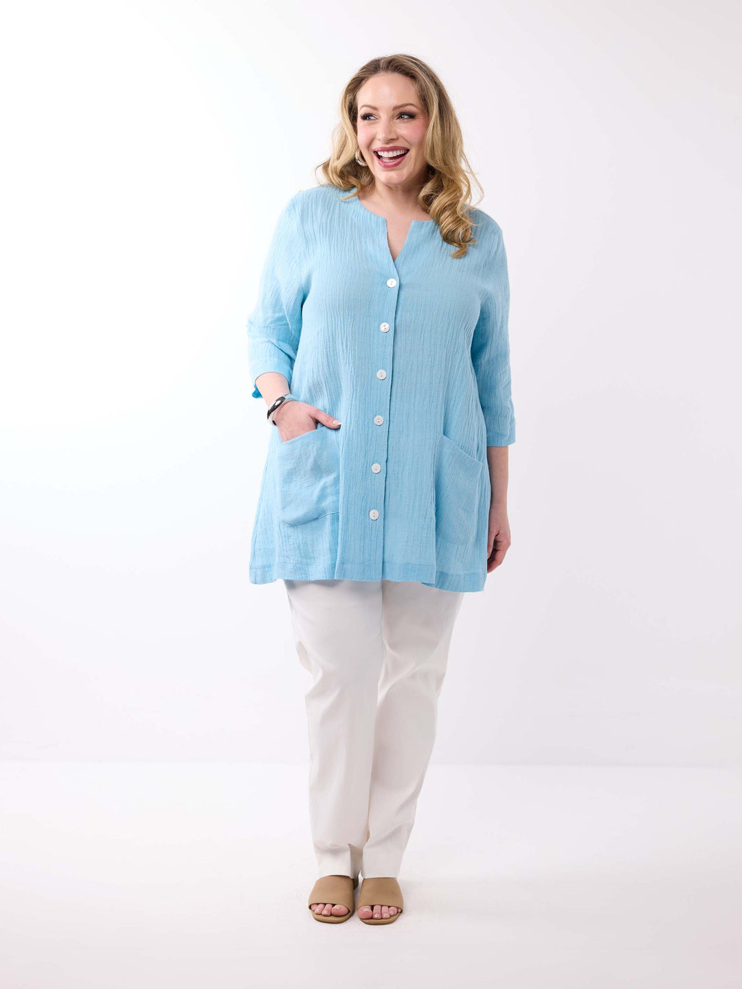 A woman wearing a light aqua blue crinkle-textured button-up top with white buttons and side pockets, paired with white pants