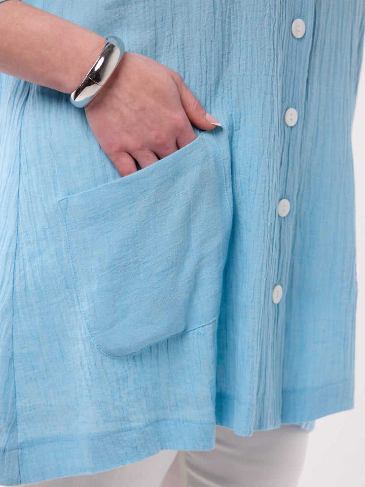 Close-up of a person wearing a light blue crinkled linen shirt with a front pocket and white buttons, showing a hand in the pocket