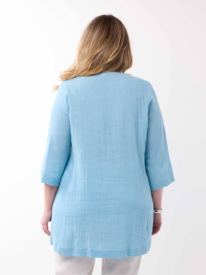 A woman viewed from the back wearing a light blue, textured three-quarter sleeve tunic top and white pants