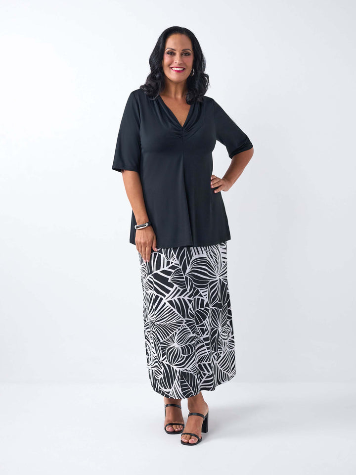 A woman wearing a black short-sleeve top with a draped cowl neckline, paired with black and white tropical palm patterned skirt