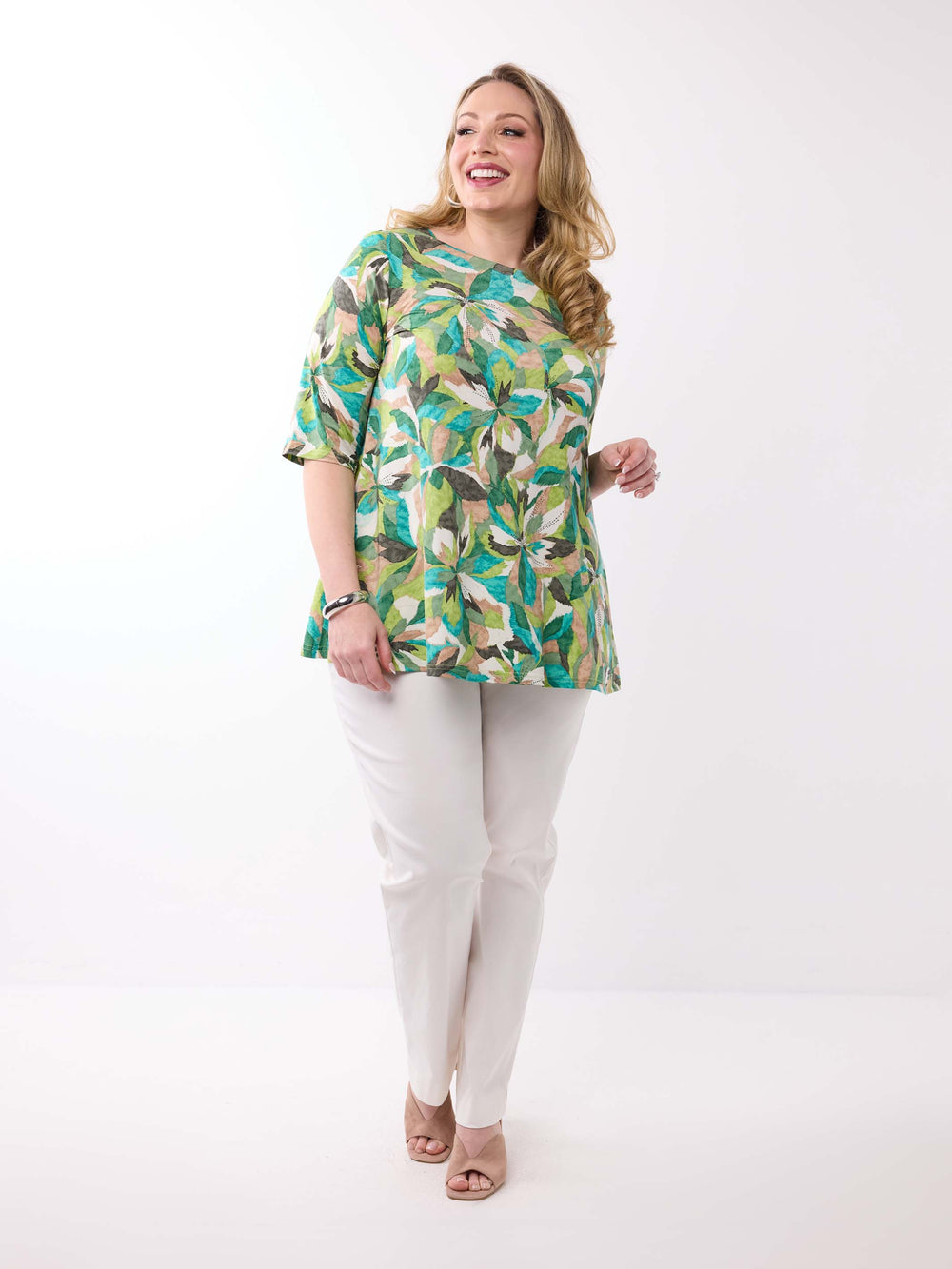 A woman wearing a short-sleeve, crew-neck top with a vibrant green tropical leaf pattern, paired with white pants