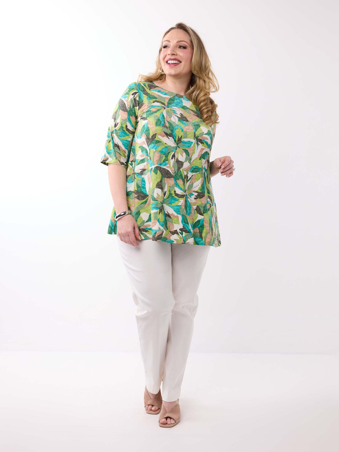 A woman wearing a short-sleeve, crew-neck top with a vibrant green tropical leaf pattern, paired with white pants