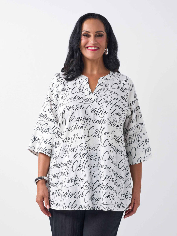 A woman wearing a white three-quarter sleeve top with an all-over black script coffee-themed print, paired with black pant