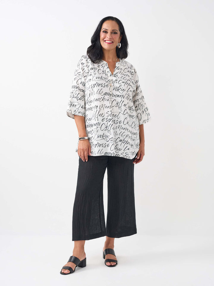 A woman wearing a white three-quarter sleeve top with an all-over black script coffee-themed print, paired with black pant