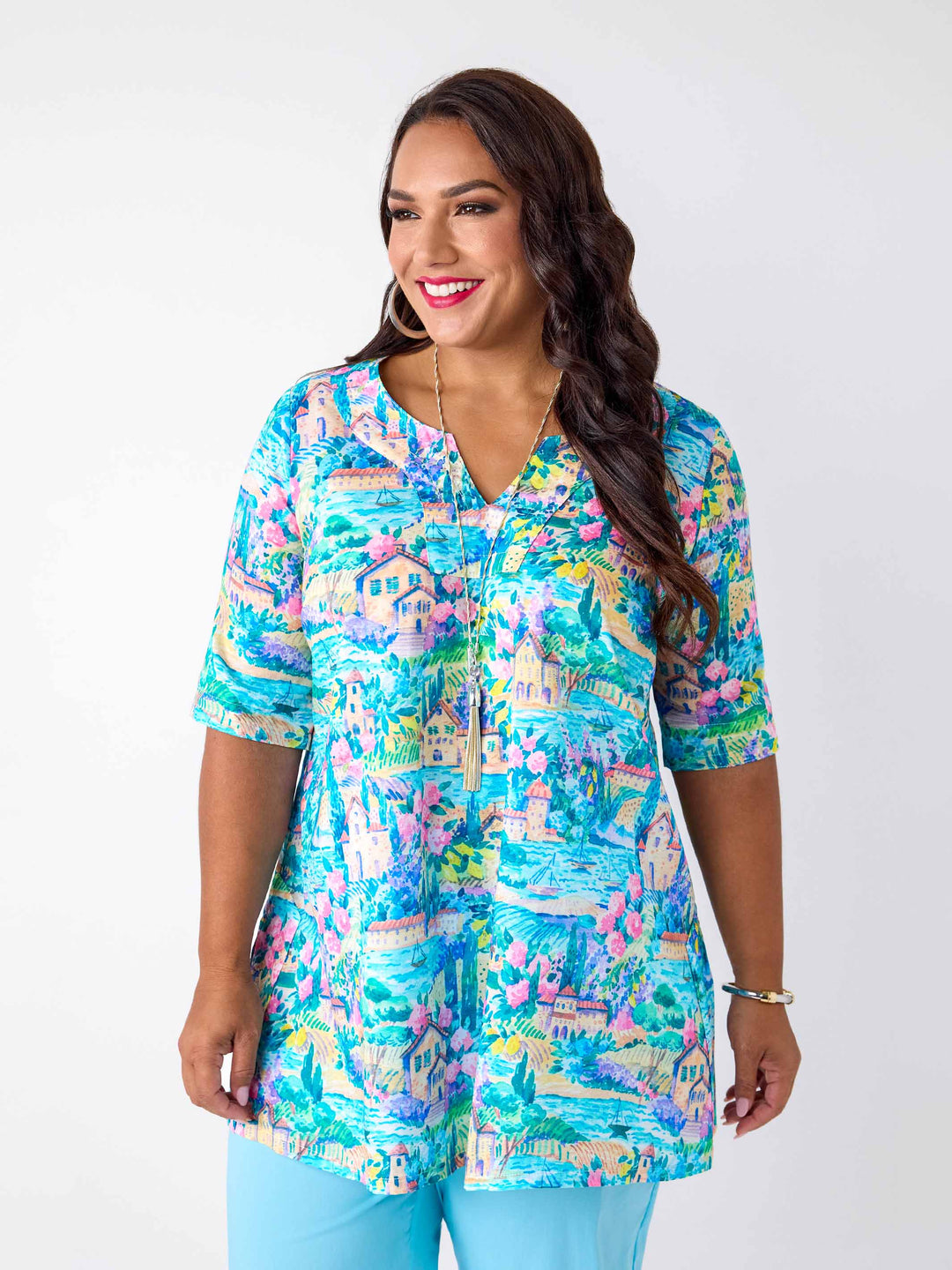 A woman wearing a short-sleeve notch neck cotton top with a colourful tropical lake print in blue and pink, paired with light blue pants