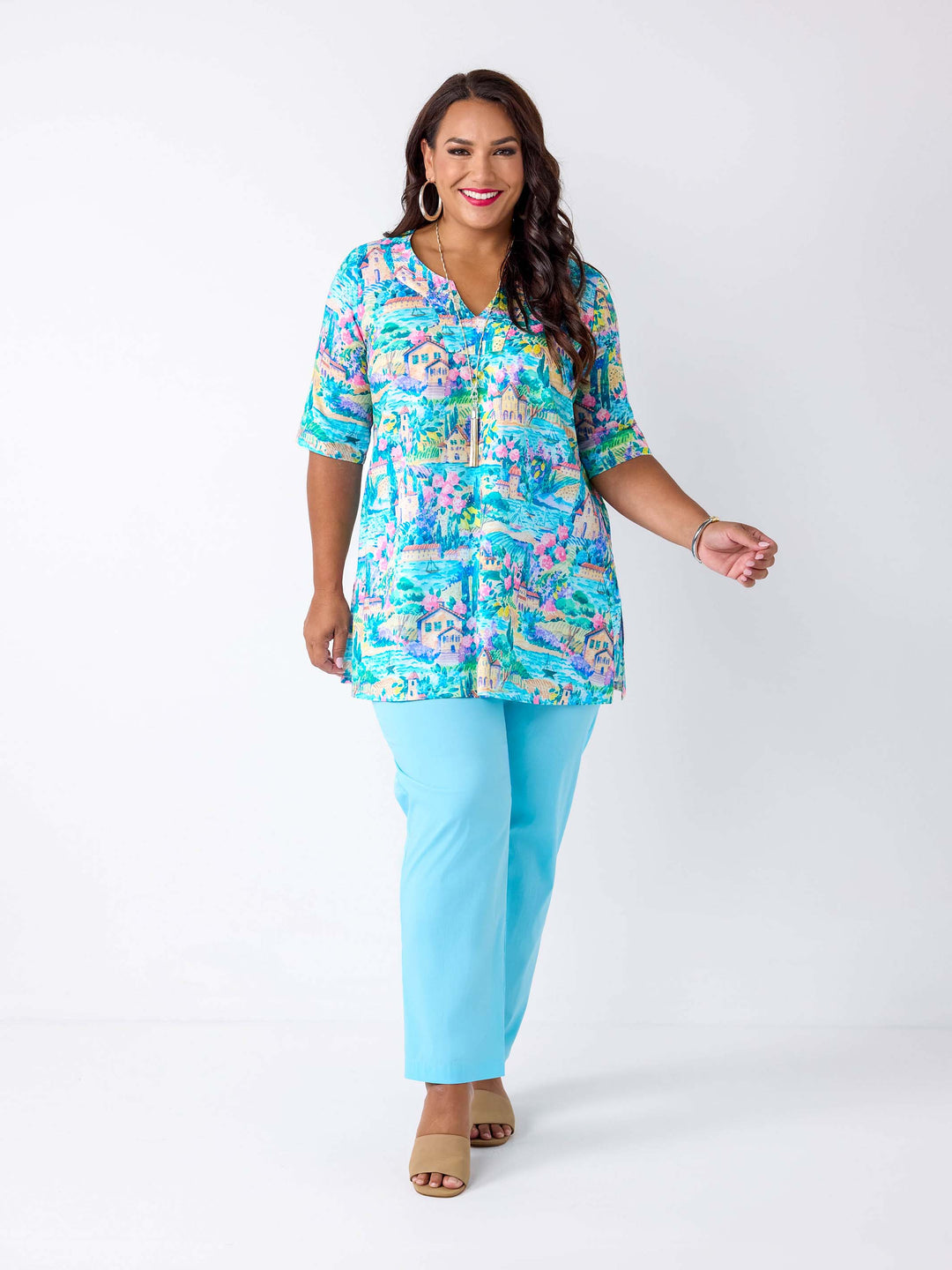 A woman wearing a short-sleeve notch neck cotton top with a colourful tropical lake print in blue and pink, paired with light blue pants