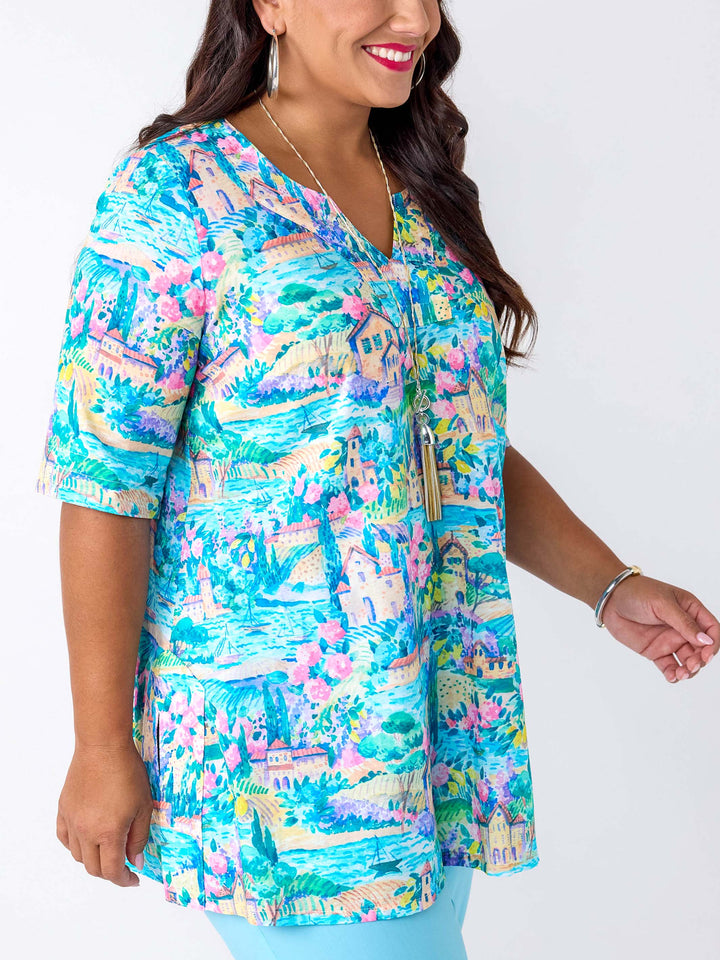 A woman wearing a short-sleeve V-neck tunic top with a colourful tropical landscape print in turquoise, pink, green, and beige, paired with light blue pants