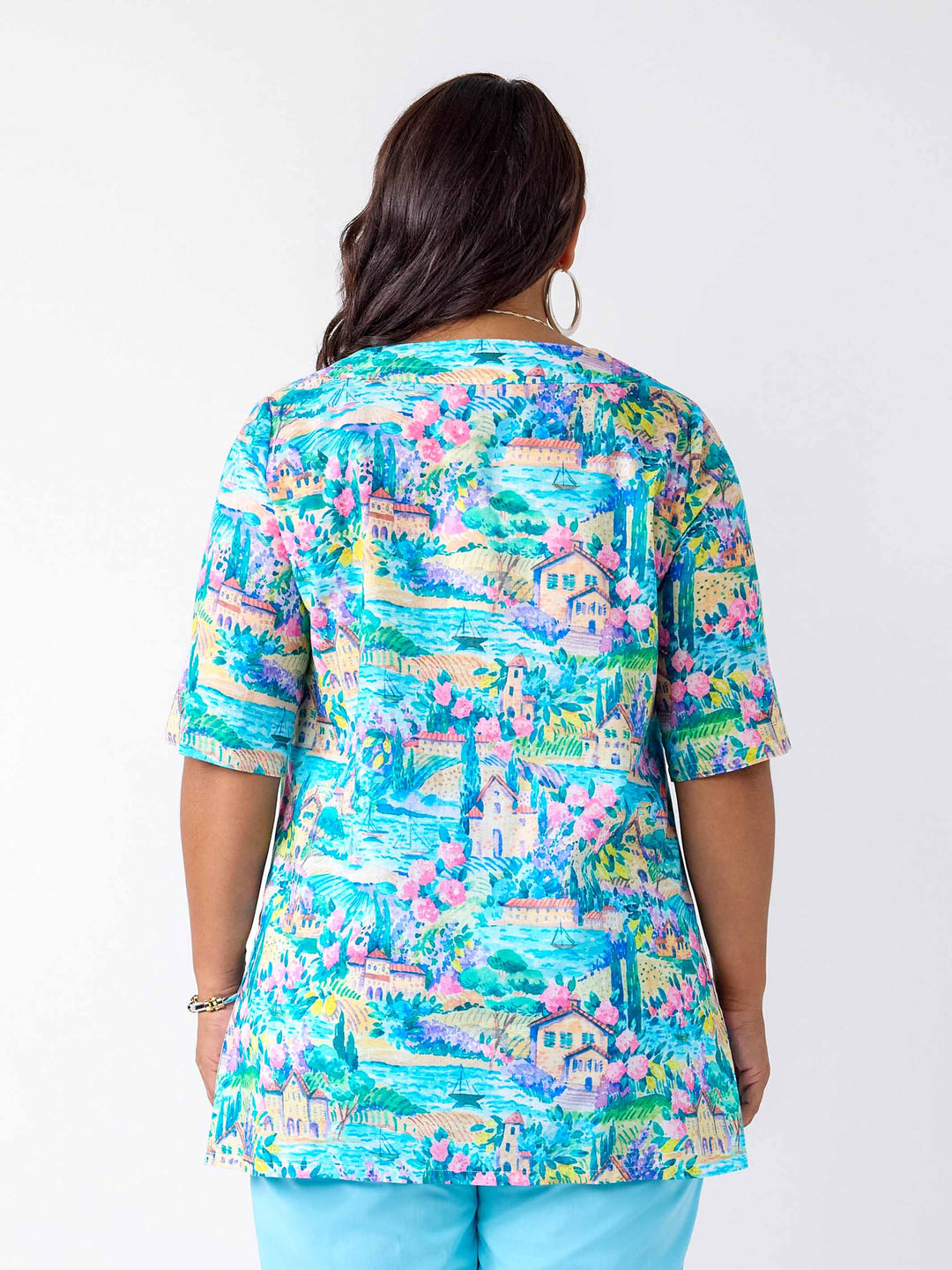 A woman shown from the back wearing a short-sleeve tunic top with a colorful, detailed landscape print featuring blue skies, green trees, pink flowers, and houses.